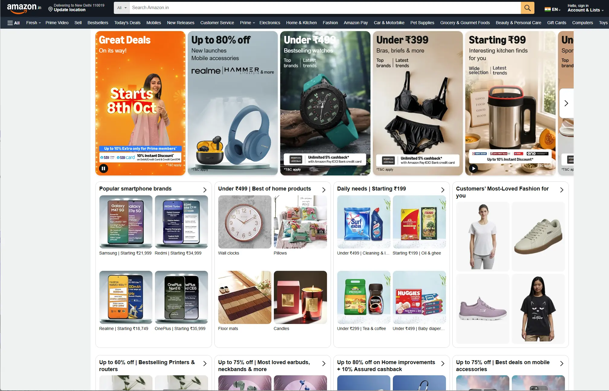 A screenshot of the Amazon India homepage featuring a top banner with deals starting on October 8th. The banner includes promotions for up to 80% off on Realme audio, watches under ₹499, bras under ₹399, and kitchen finds starting at ₹99. Below the banner, four product category sections are visible: "Popular smartphone brands" showing Samsung, Redmi, Realme, and OnePlus phones; "Under ₹499 | Best of home products" showing wall clocks, pillows, floor mats, and candles; "Daily needs" showing detergents, oils, tea, and baby diapers; and "Customers' Most-Loved Fashion" displaying t-shirts and sneakers.
