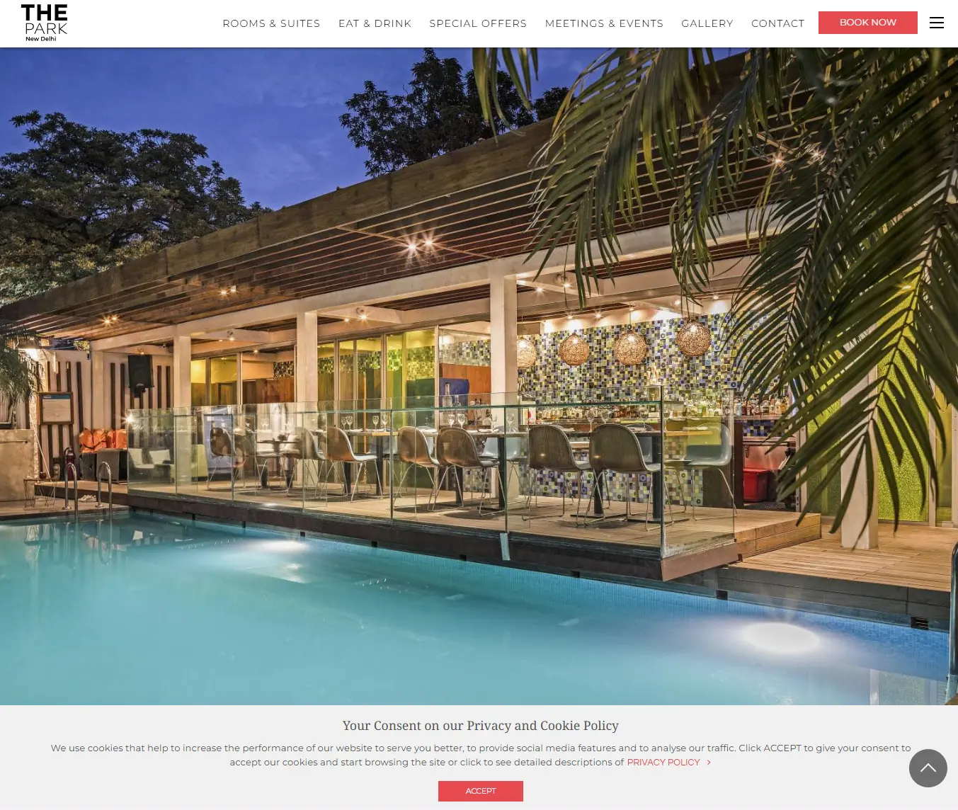 Outdoor poolside restaurant view at The Park Hotel, New Delhi, showcasing modern glass architecture, elegant dining seating, mosaic-tile bar counter, and warm ambient lighting under a wooden pergola.